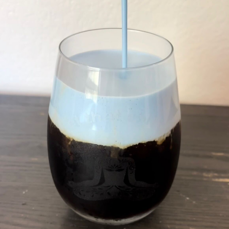 Close up of glass of low-sugar blue cold brew showcasing pale blue cold foam being poured on top with wooden table background