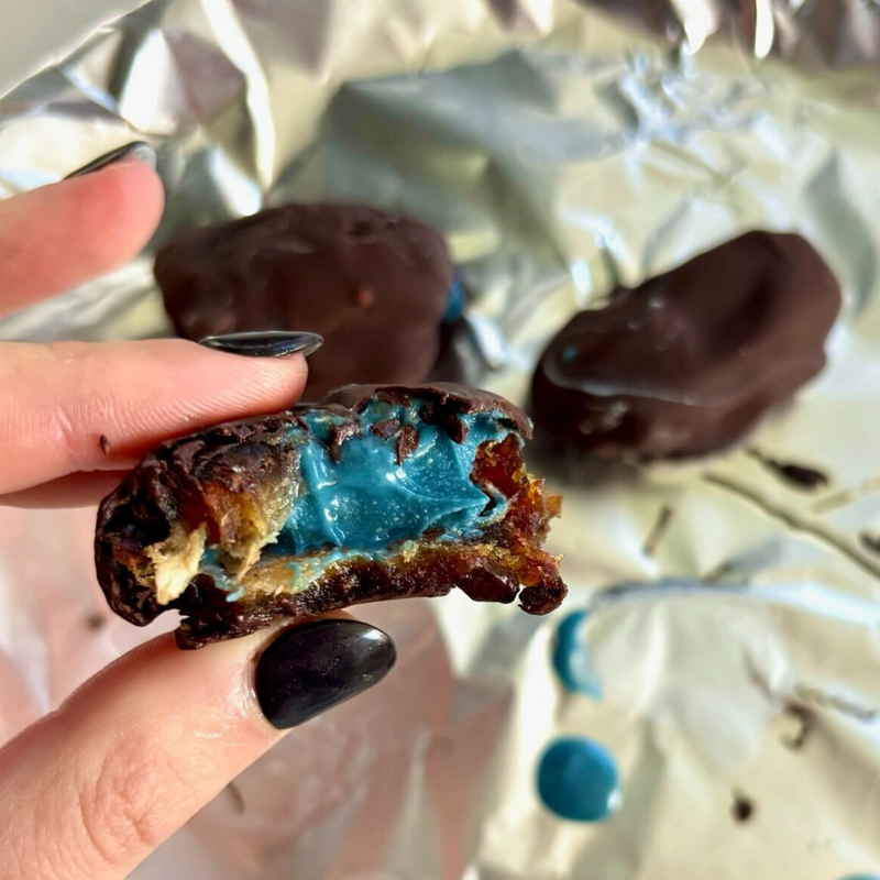 Close-up of a chocolate-coated date bitten to show bright blue cashew butter filling, held over crinkled foil