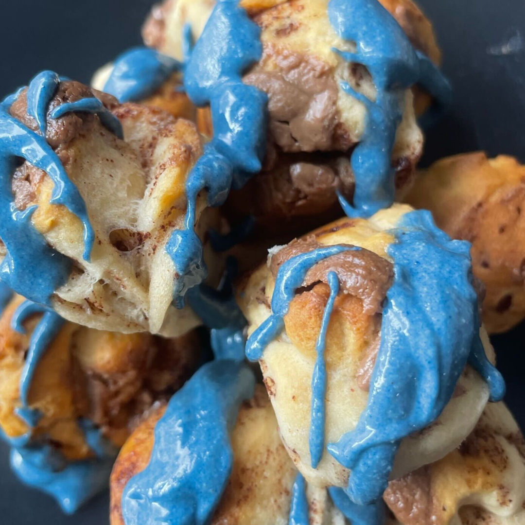 Close-up of blue cinnamon roll mini donuts showing blue spirulina vanilla and chocolate fudge cashew butter drizzles on a dark plate