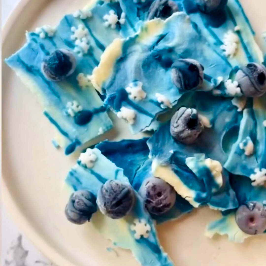 Overhead view of blue yogurt bark showcasing blue and white swirls, frozen blueberries, and snowflake sprinkles on a white plate
