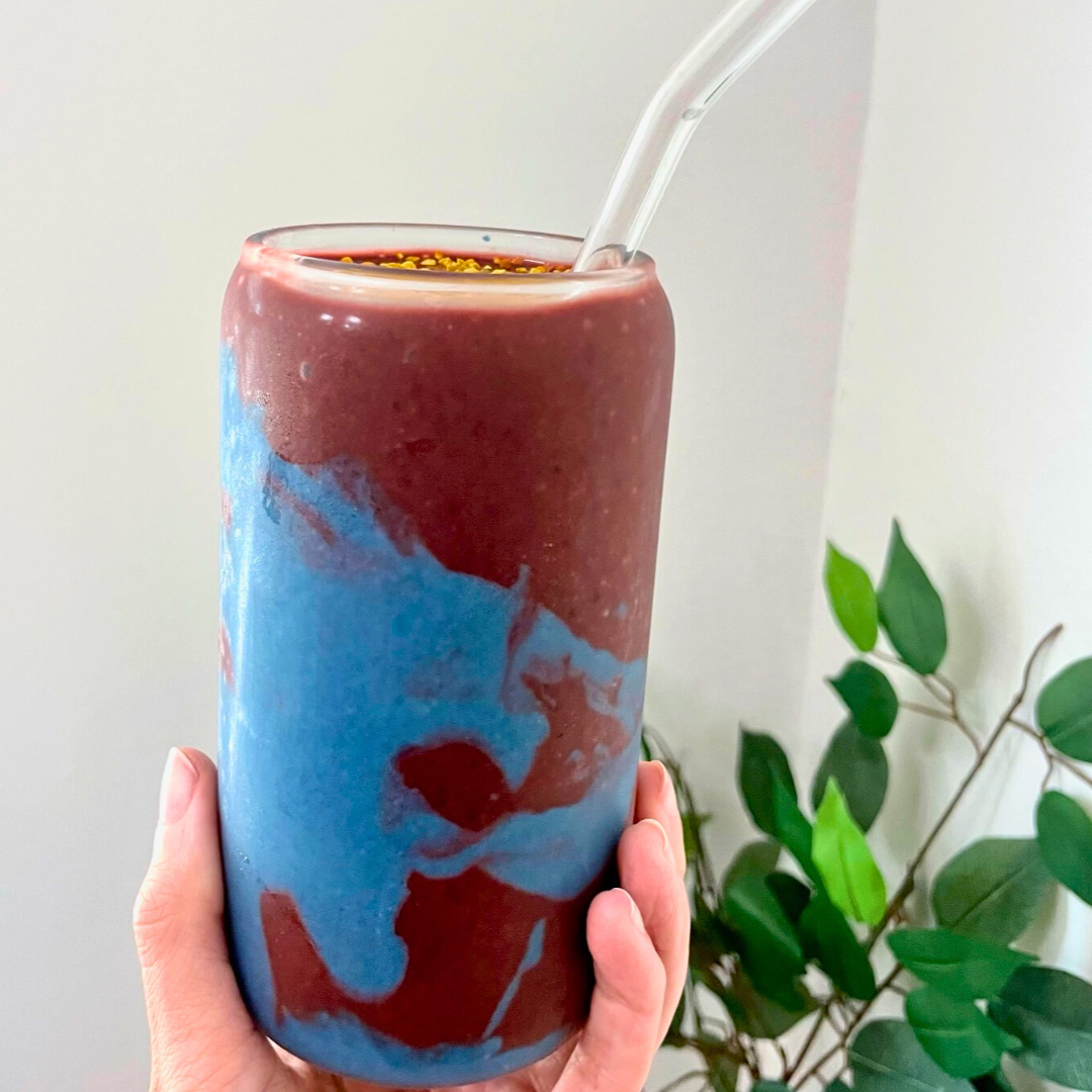 Hand holding tall glass with marbled blue and raspberry-red smoothie topped with bee pollen and a clear straw, plant in background