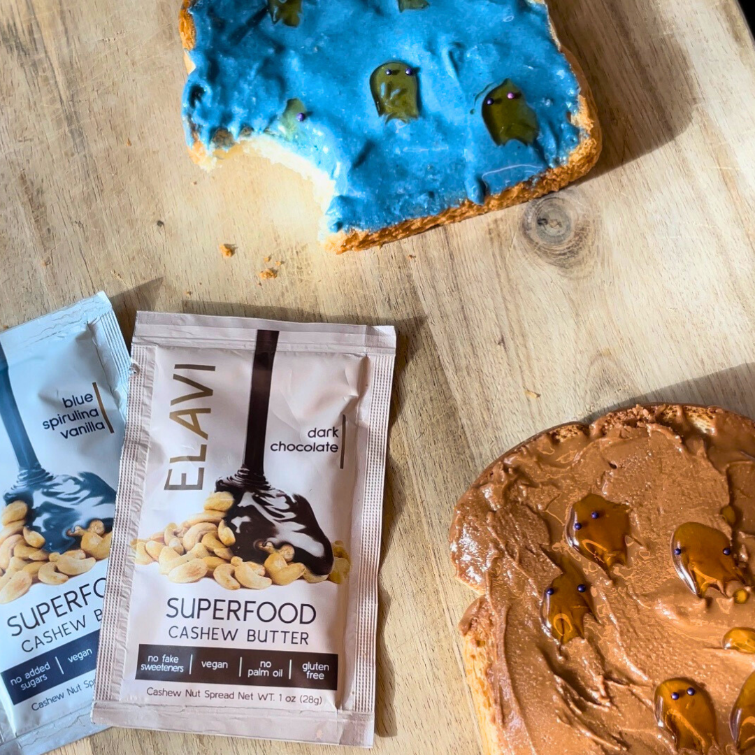 Two slices of ‘ghost’ toast — one with blue frosting and a bite taken, one with chocolate cashew butter — arranged with ELAVI single-serve cashew butter packets on a wooden board