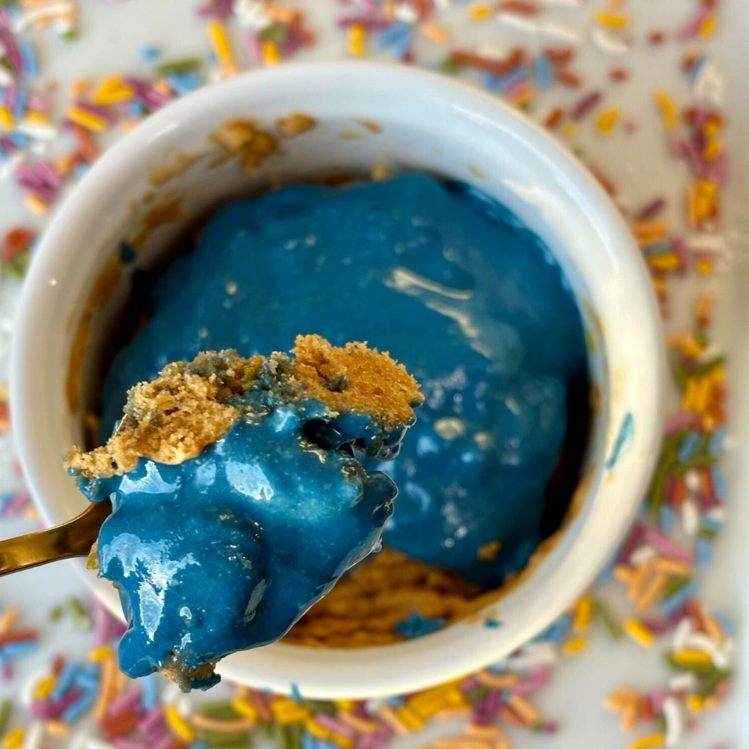 Vegan funfetti mug cake topped with bright blue ELAVI cashew butter frosting and rainbow sprinkles on a white plate