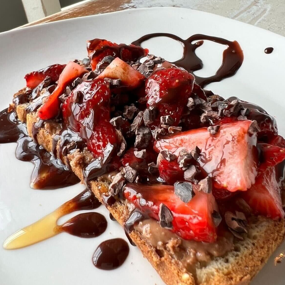 Toast topped with ELAVI Chocolate Fudge Cashew Butter, sliced strawberries, cacao nibs, and dark chocolate drizzle on a white plate