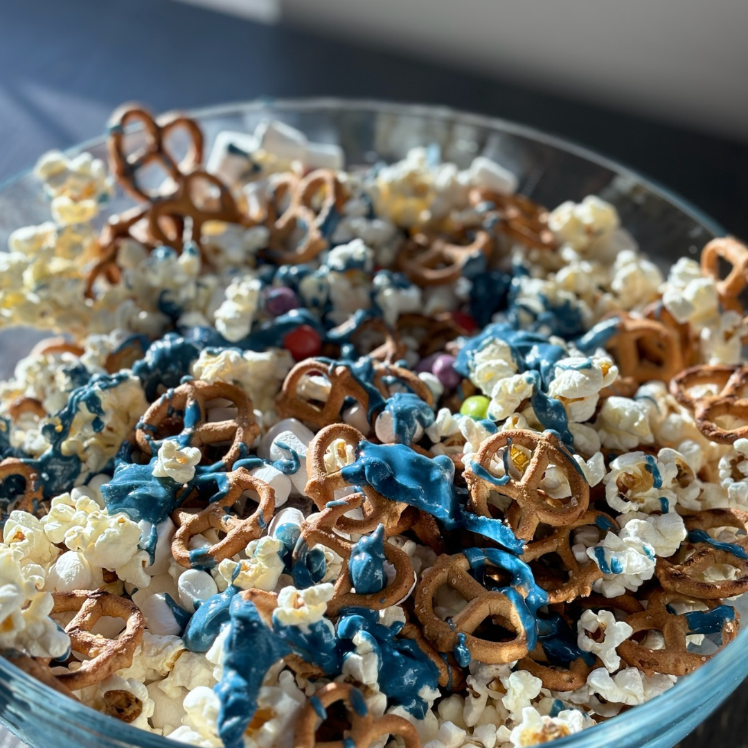 Close-up of Sweet & Salty popcorn mix showing popcorn, mini pretzels, marshmallows, candy pieces and blue frosting drizzle in a glass bowl