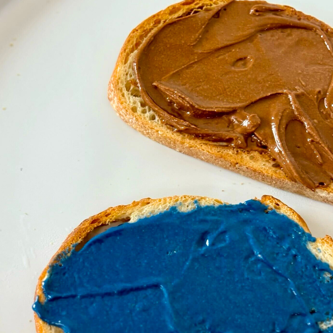 Two slices of toasted bread on a white plate, one spread with chocolate nut butter and the other with a vibrant blue spread