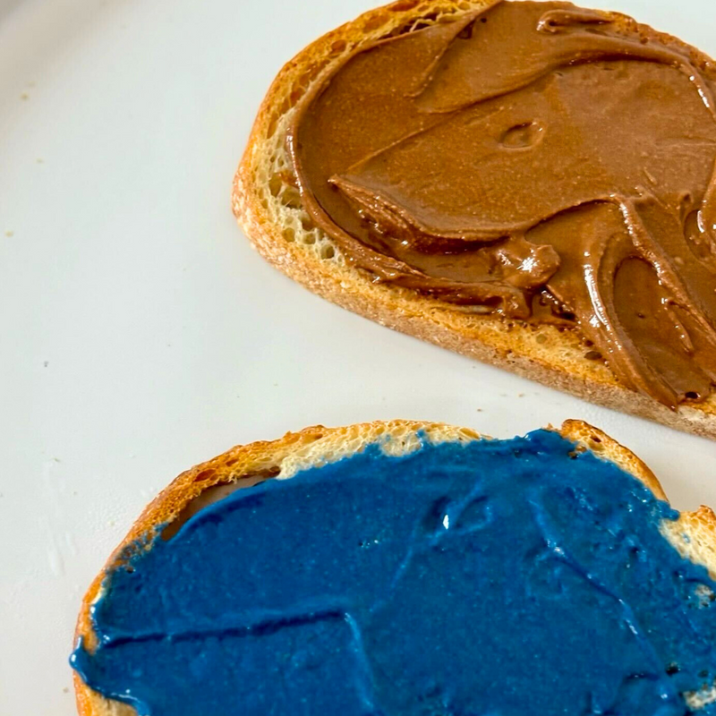 Two slices of toasted bread on a white plate, one spread with chocolate nut butter and the other with a vibrant blue spread