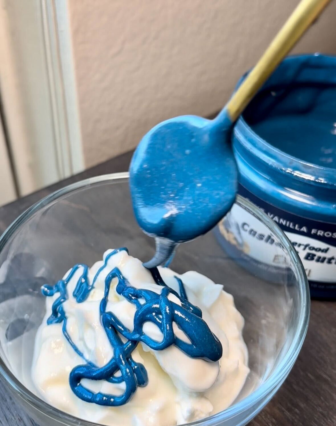 Spoonful of bright blue spirulina drizzling onto a bowl of yogurt, jar of blue cashew butter at right