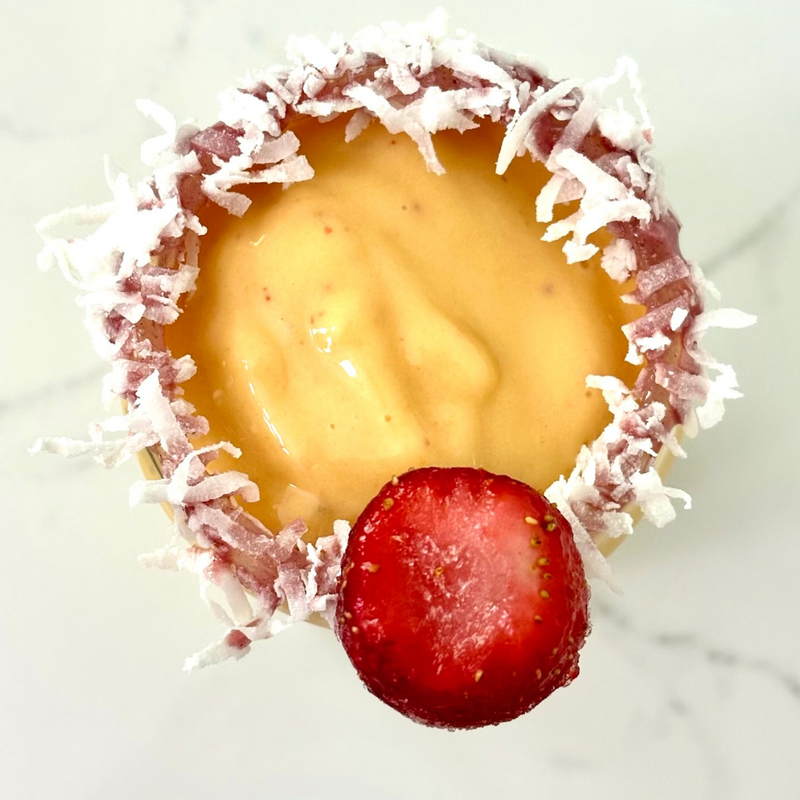 Top view of a strawberry mango smoothie in a glass with shredded coconut rim and a halved strawberry garnish on a white marble surface