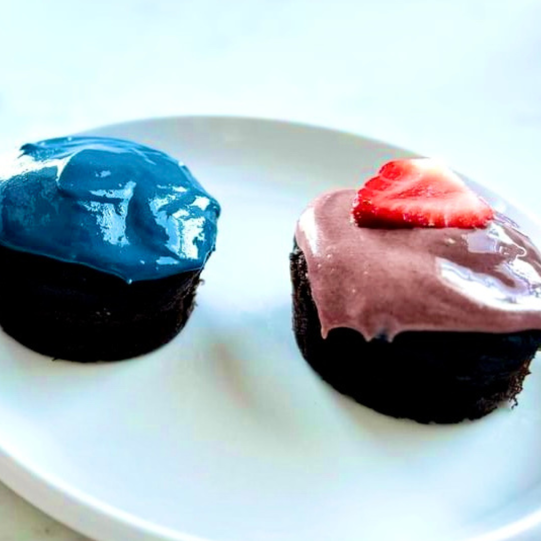 Top view of mini healthy chocolate cake cupcakes with blue and pink frosting, one topped with strawberry on white plate
