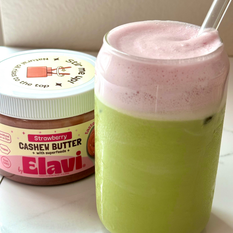 Close-up of iced matcha in glass with pink strawberry cold foam, beside Elavi Strawberry Cashew Butter jar on white surface