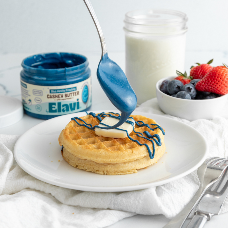 Waffle with butter pat while blue Elavi cashew butter is drizzled from a spoon, jar of milk and berries behind