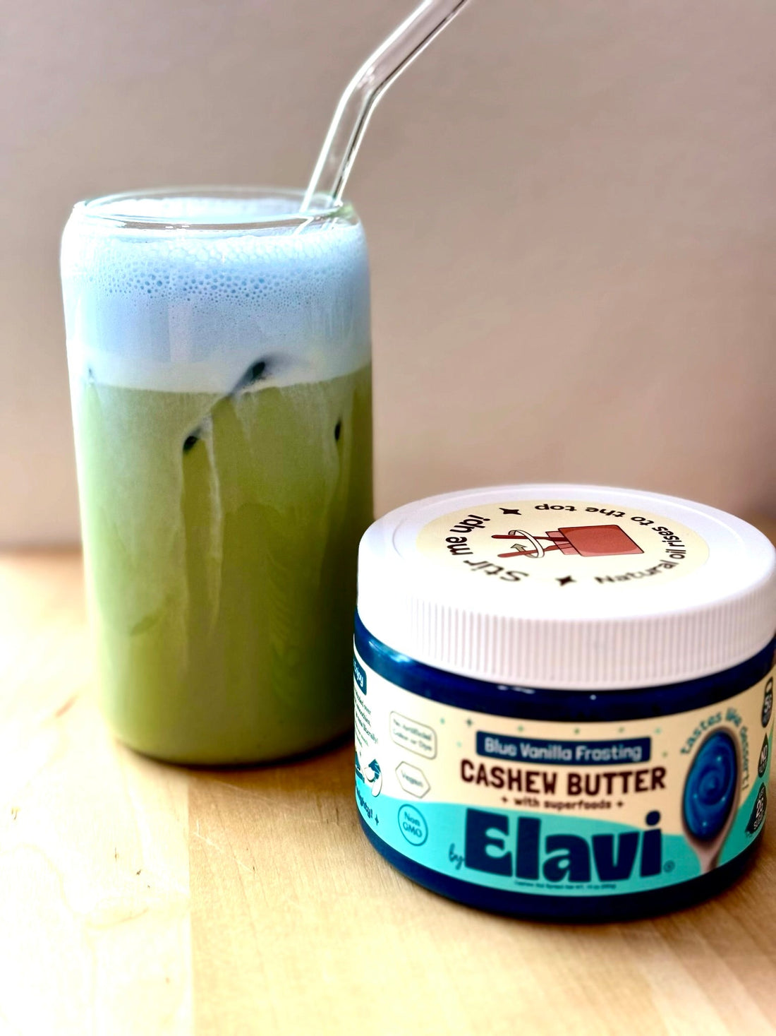 Iced matcha topped with blue vanilla froth beside an Elavi Blue Vanilla Frosting cashew butter jar on a wooden table