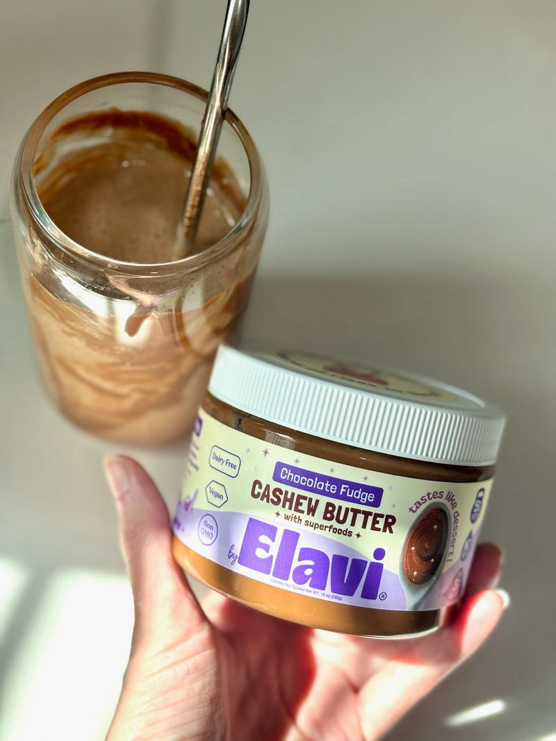 Angled close-up of Elavi Chocolate Fudge Cashew Butter jar held in hand with blended chocolate drink and spoon in background