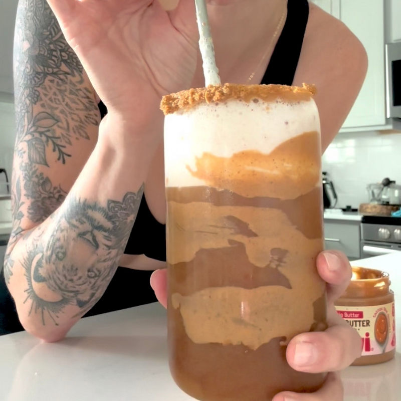 Person holding a layered protein cookie-butter cold brew latte with frothy milk and cookie-crumb rim in a kitchen, Elavi Cookie Butter jar visible