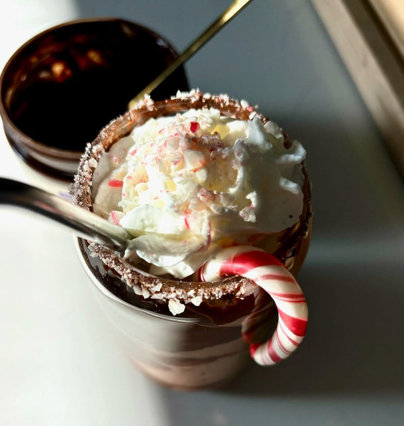 Peppermint cocoa protein smoothie in a glass topped with whipped cream and crushed peppermint, chocolate-rimmed glass with a mini candy cane and spoon