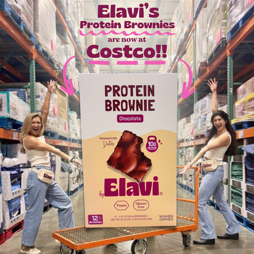 Two women pose with a giant Elavi protein brownie box in a Costco warehouse; overlay text reads Elavi’s Protein Brownies are now at Costco