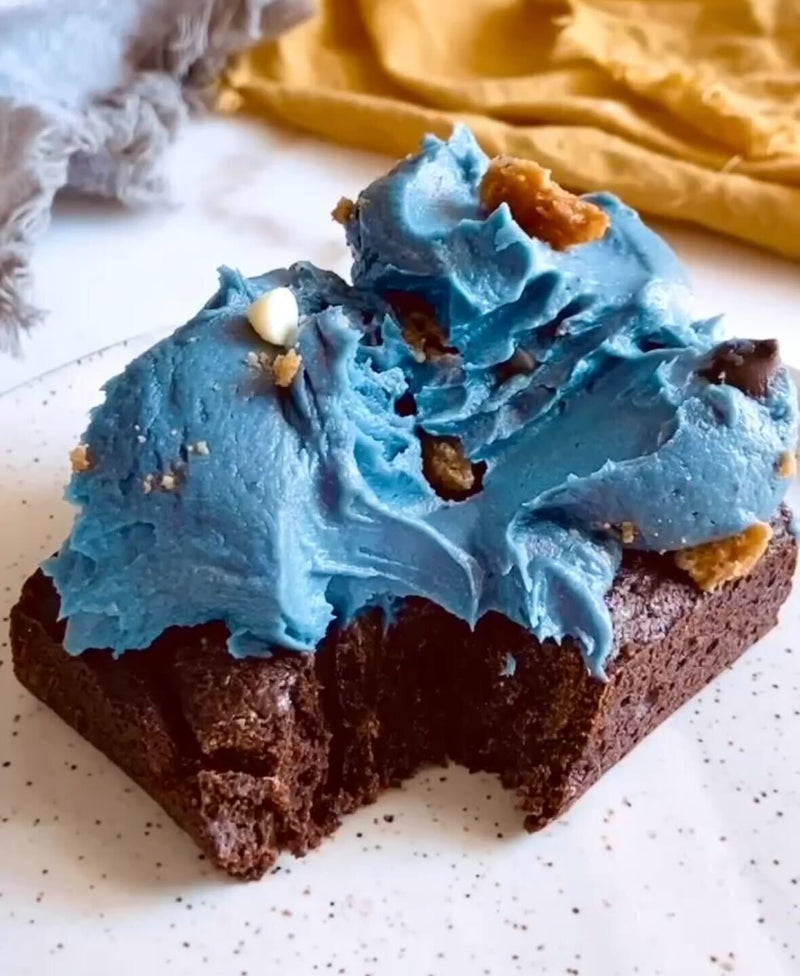 Close-up of brownie topped with bright blue whipped frosting showcasing fluffy swirls and cookie crumbs on a speckled plate