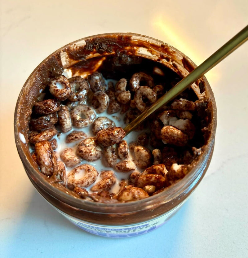 Almost empty jar of chocolate fudge cashew butter used as a cereal bowl with cereal, milk, and a spoon on a light countertop