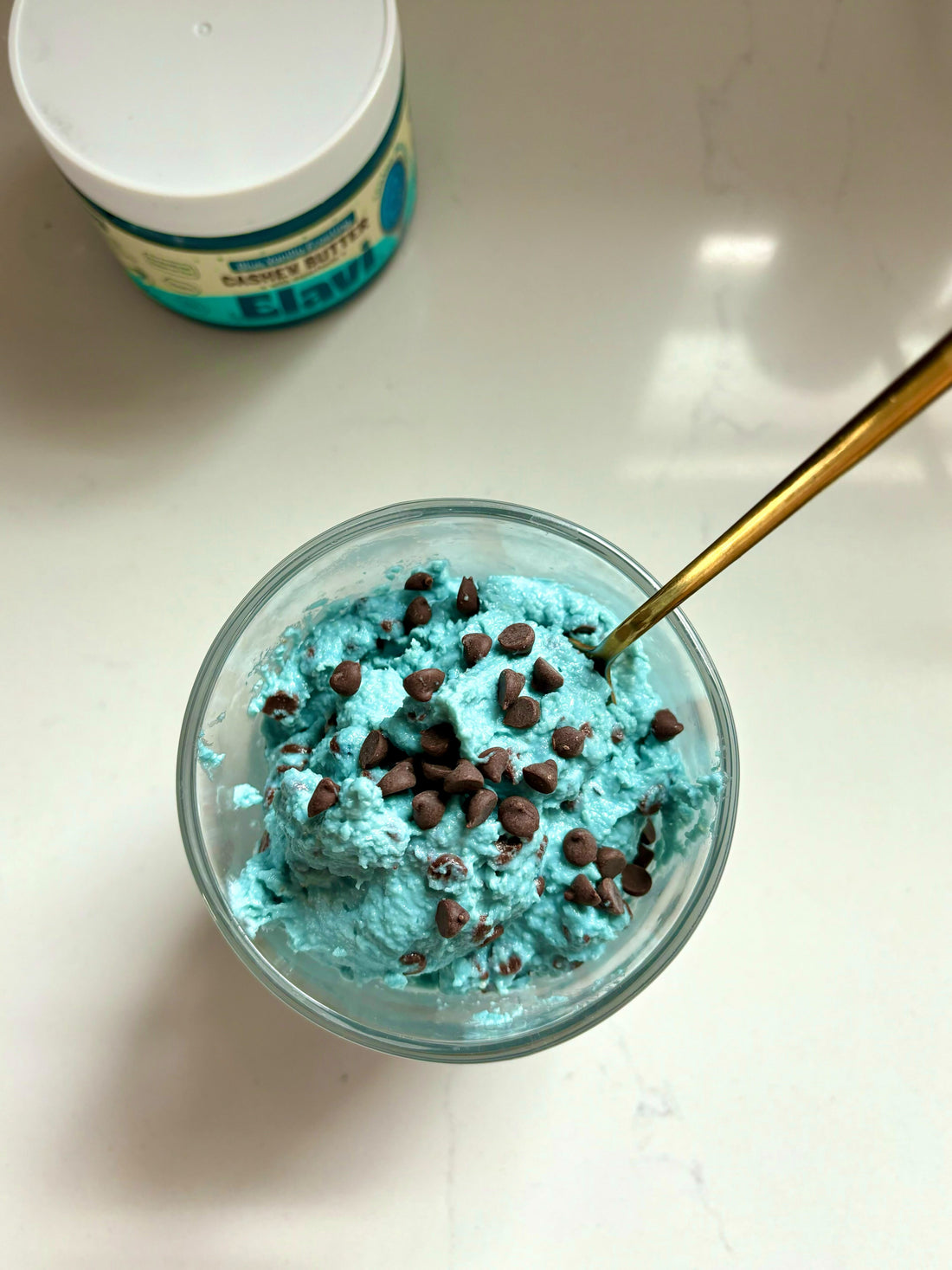 Top-down view of bright blue cottage-cheese cookie dough with chocolate chips in a glass bowl and a gold spoon on a white countertop