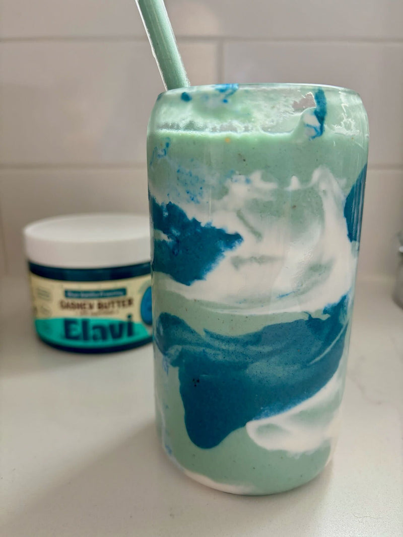 Front view of coconut cloud smoothie in a tall glass with blue and white swirls and a straw, a jar blurred in the background