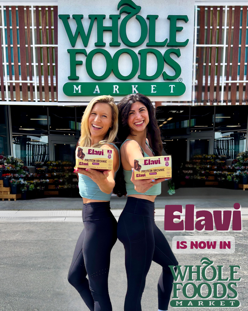 Two women holding Elavi Protein Brownie boxes outside a Whole Foods Market entrance with overlaid text Elavi is now in Whole Foods Market