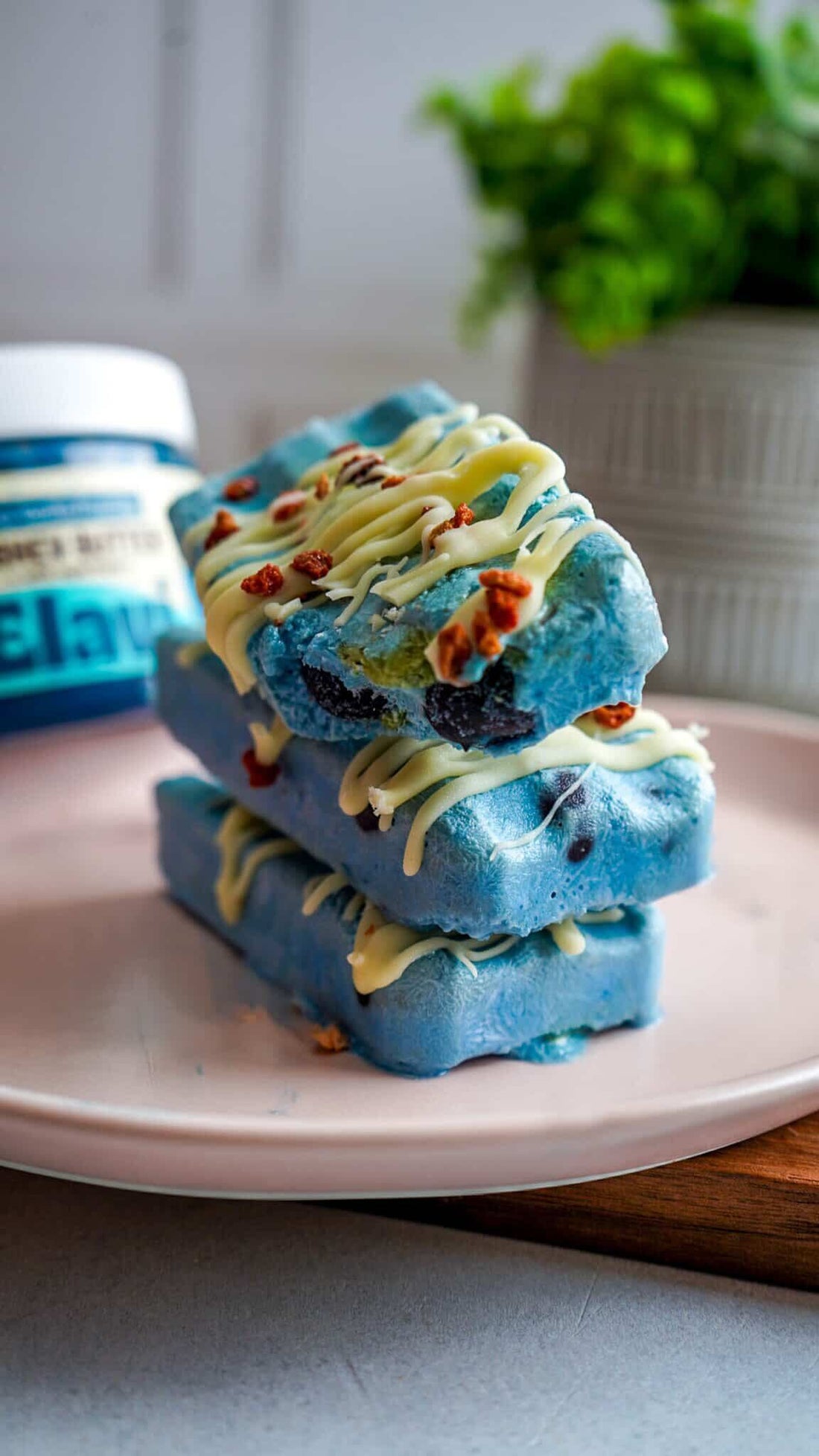Close-up of blueberry matcha popsicles drizzled with white chocolate and chopped nuts, stacked on a pink plate with a blurred jar behind
