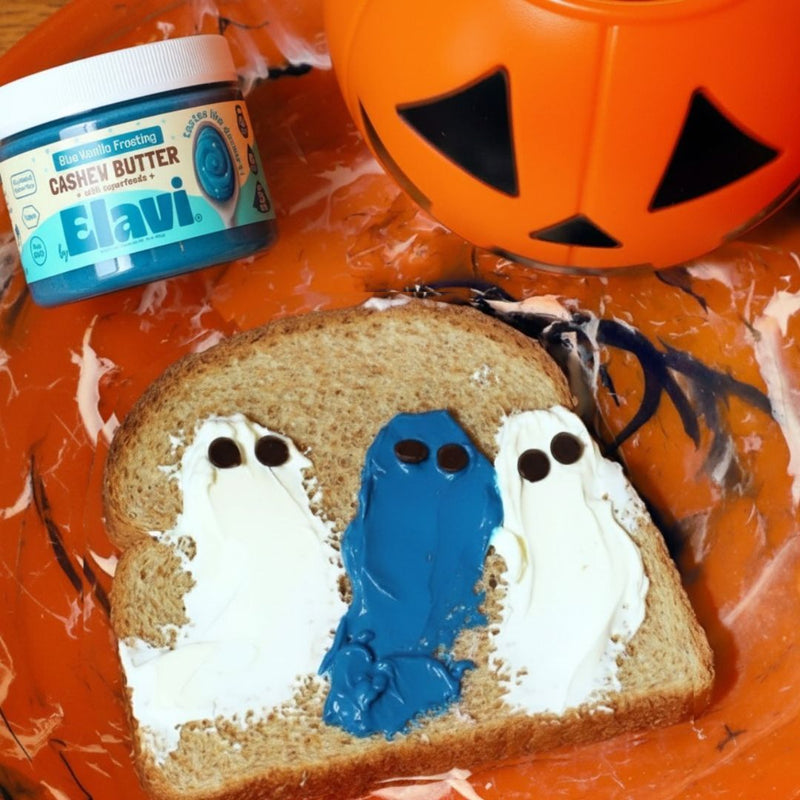 Toast with three ghost-shaped spreads of white and blue Elavi cashew butter frosting on an orange Halloween plate
