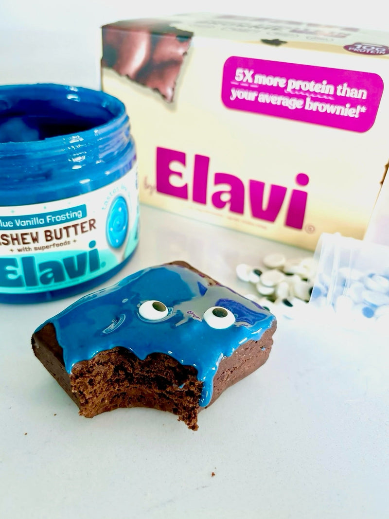Close-up of Elavi Blue Monster protein brownie with blue frosting and edible googly eyes, bitten, jar and box in background