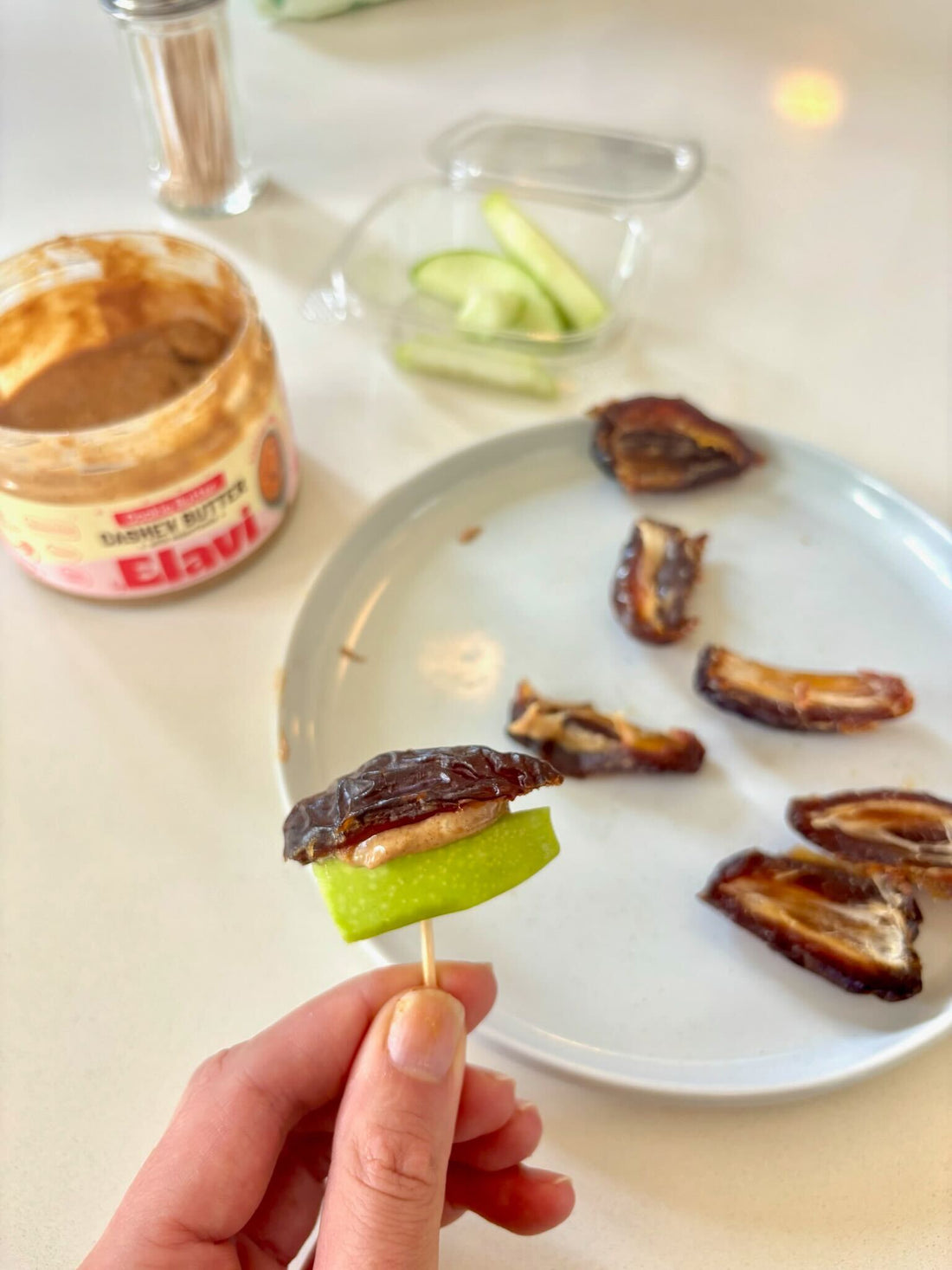 Hand holding toothpick with green apple slice, nut butter, and halved date; plate and jar of Elavi nut butter in background