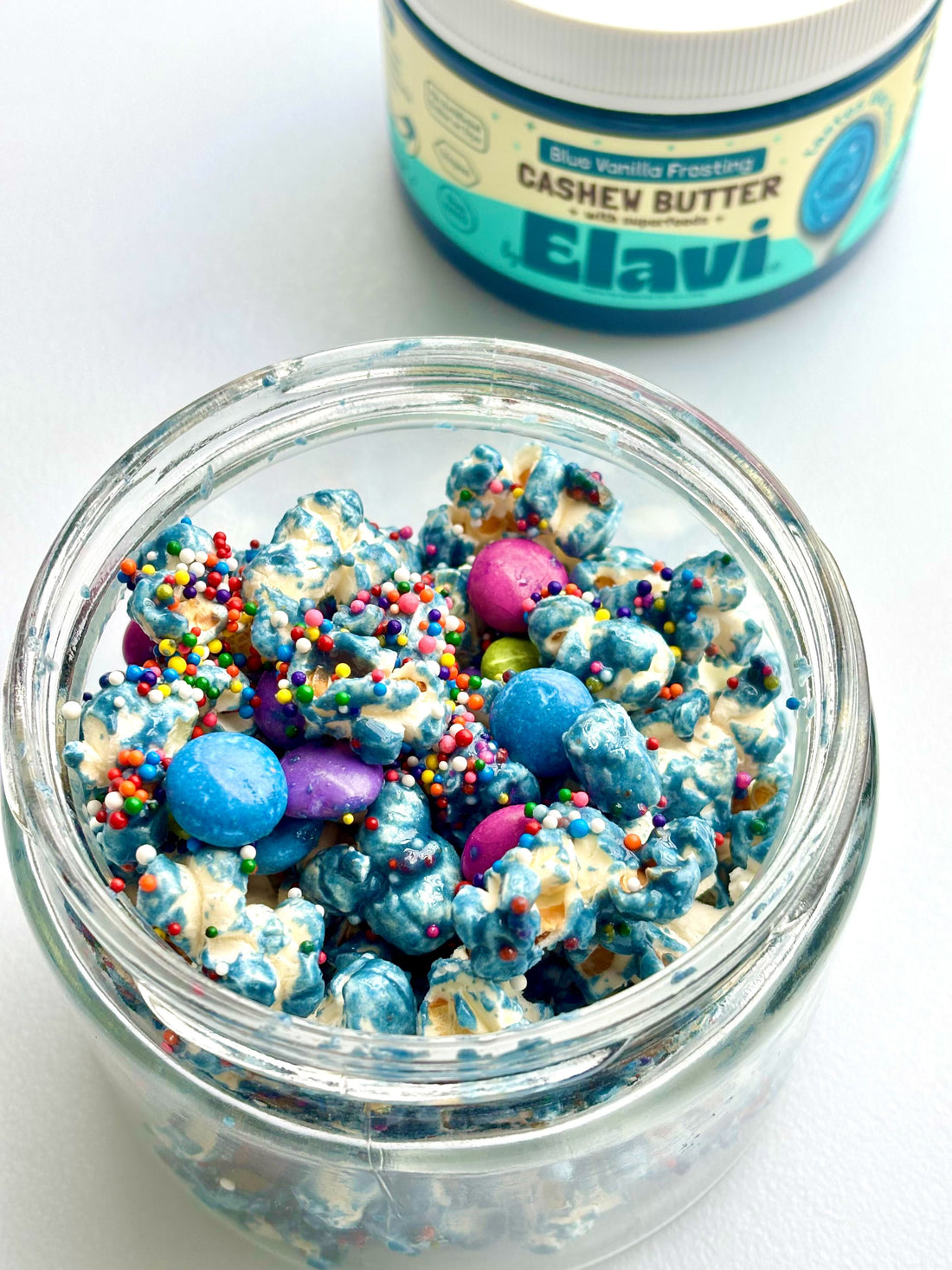 Top-down view of unicorn popcorn in a jar with blue swirled coating, sprinkles and candy gems, Elavi cashew butter jar behind