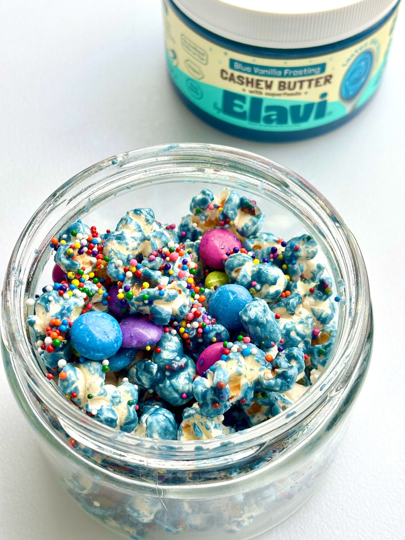 Top-down view of unicorn popcorn in a jar with blue swirled coating, sprinkles and candy gems, Elavi cashew butter jar behind