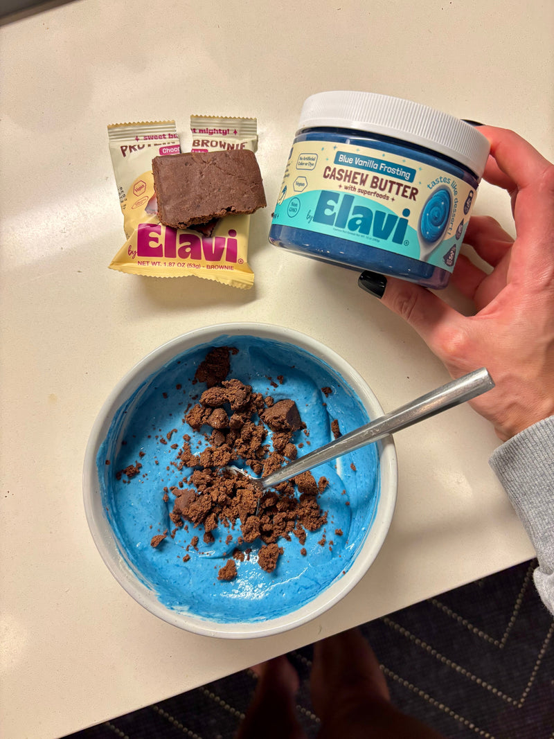 Bowl of bright blue protein pudding topped with chocolate brownie pieces, spoon in bowl, Elavi cashew butter jar and brownies nearby