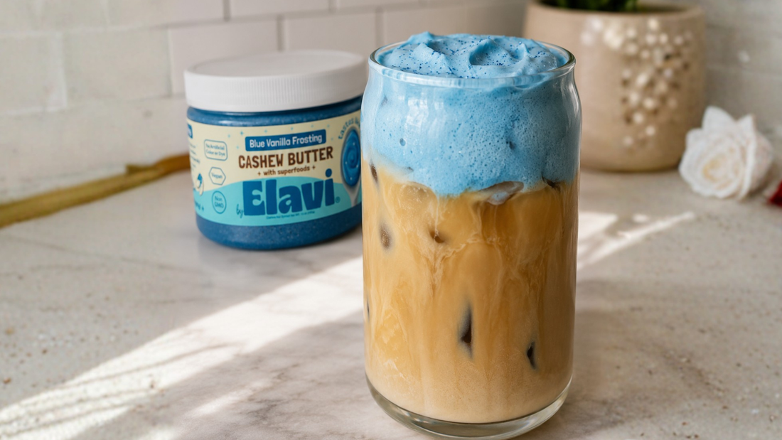 Iced coffee topped with blue protein cold foam beside a jar of Elavi Blue Vanilla Frosting cashew butter on a marble countertop