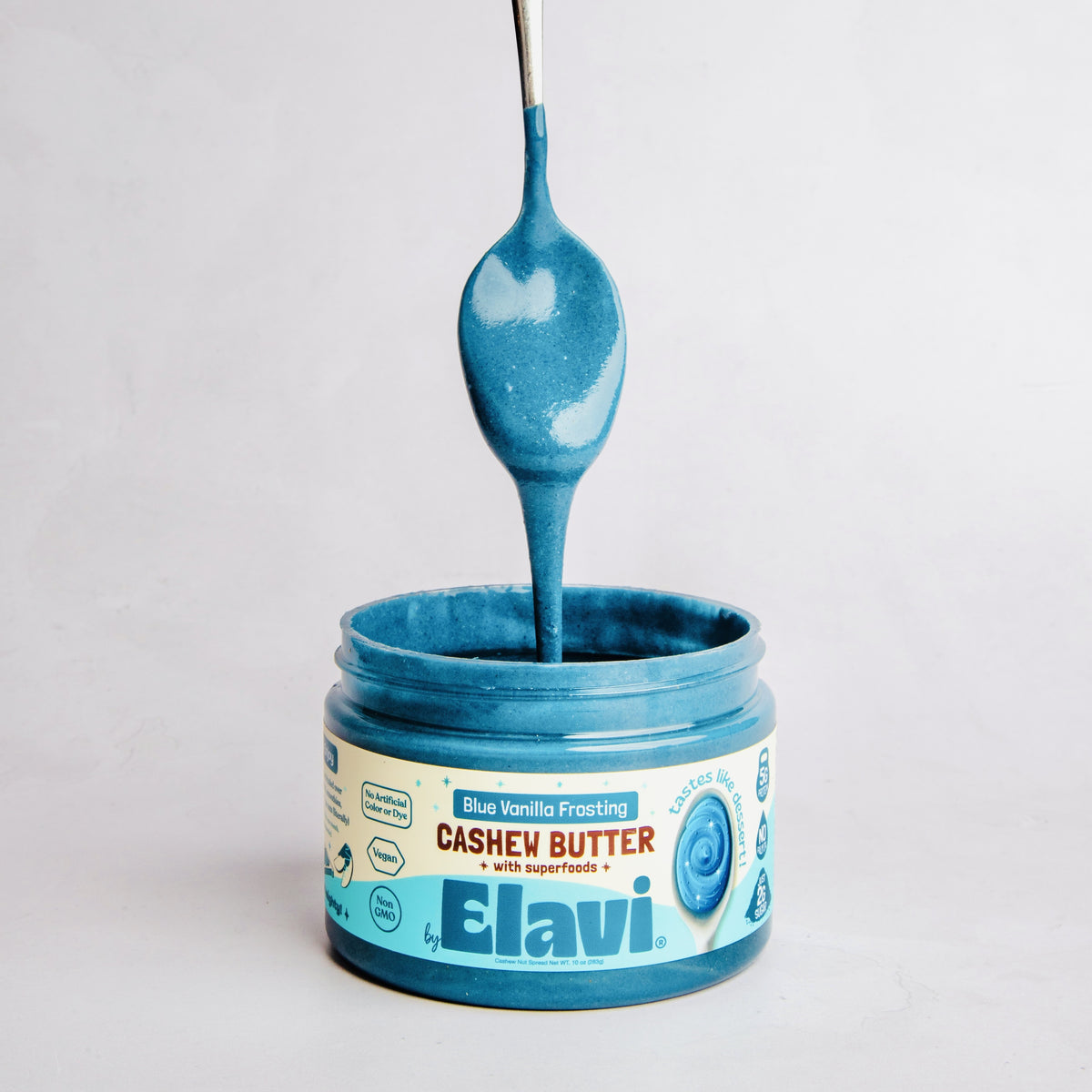 Front of Blue Vanilla Frosting cashew butter jar with spoon dripping blue frosting and Elavi branding on light gray