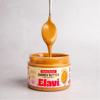 Front view of open jar of Elavi Cookie Butter cashew butter with spoon drizzling creamy spread on a light background