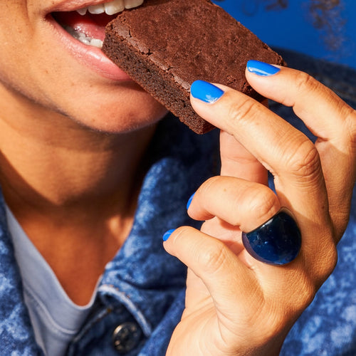Person biting a chocolate brownie while holding it with bright blue painted nails against a denim jacket background